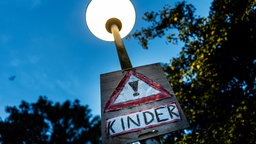ymbolbild: Nahaufnahme einer Straßenlaterne, an deren Mast ein warnendes Schild mit der Aufschrift "Kinder" angebracht ist.