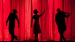 Die Schatten dreier Schauspielenden sind auf einem roten Theatervorhang zu sehen