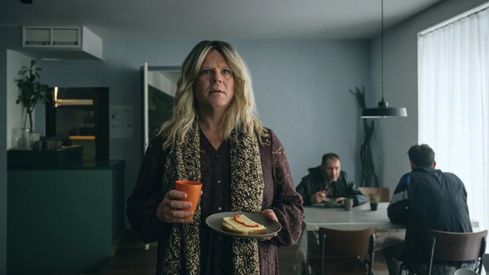 Ein Darsteller der Serie "Toxic Tom" steht kostümiert als Frau mit einem Becher und einem Teller in den Händen in einem Raum.