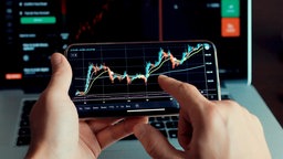 Nahaufnahme zweier Hände die ein Smartphone halten, darauf zusehen ist die grafische Darstellung einer Aktienmarktanalyse mit Kursdaten und Preischarts.