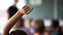Ein Kind zeigt im Unterricht auf – Nahaufnahme der Hand und des Arms, im hintergrund vershcommen der Lehrer und die Klasse.