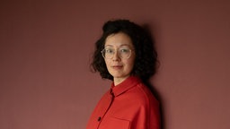 Das Foto zeigt Yuko Kuhn mit offenem, dunklem, lockigem Haar, einer Brille und einem freundlichen Lächeln.