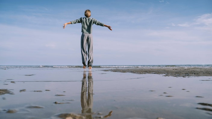 Eine Frau steht mit ausgebreiteten Armen am Strand und spiegel sich im Wasser