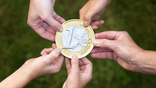Eine übergroße Euro-Münze wird von mehreren Händen gehalten