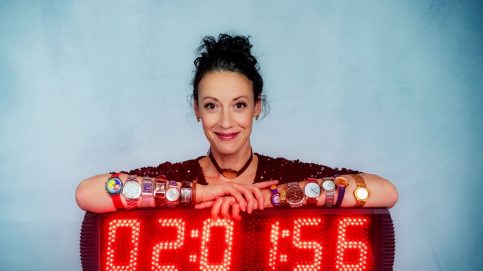 Alice Köfer stützt sich lächelnd auf einer großen Digitaluhr auf und trägt zahlreiche Armbanduhren an beiden Armen