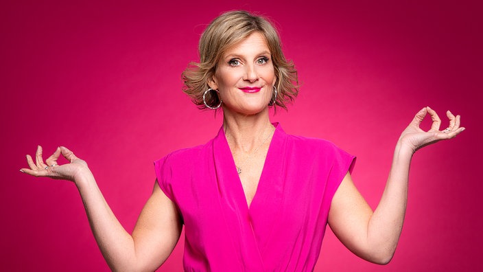 Die Comedienne Vera Deckers steht vor einem rötlich-pinken Hintergrund, trägt ein pinkes Kostüm und lächelt in die Kamera.