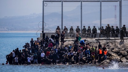 Migrant:innen warten am Grenzzaun von Ceuta auf die Überquerung nach Spanien. Dahinter steht schwer bewaffnetes Militär bereit.