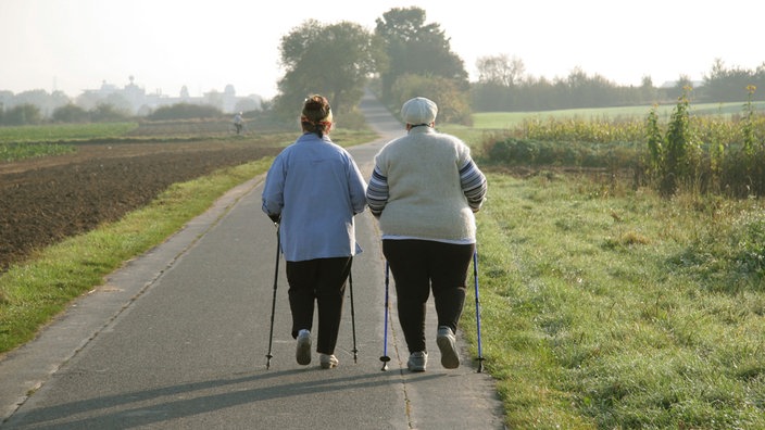 Zwei ältere Damen beim Gehen mit Skistöcken (Nordic Walking) auf einem Feldweg bei Bonn im fahlen Licht der schon tiefstehenden Sonne.