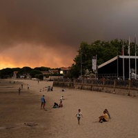 Menschen sitzen an einem Strand, im Hintergrund brennen die Wälder.