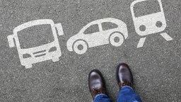 Eine weiße Bodenmarkierung auf Asphalt zeigt drei Verkehrsmittel – Bus, Auto und Straßenbahn. Unten im Bild stehen die braunen Schuhe einer Person in Jeans.