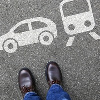 Eine weiße Bodenmarkierung auf Asphalt zeigt drei Verkehrsmittel – Bus, Auto und Straßenbahn. Unten im Bild stehen die braunen Schuhe einer Person in Jeans.