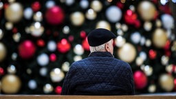 in Mann sitzt allein auf einer Bank vor einem geschmueckten Weihnachtsbaum.