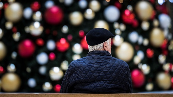 in Mann sitzt allein auf einer Bank vor einem geschmueckten Weihnachtsbaum.