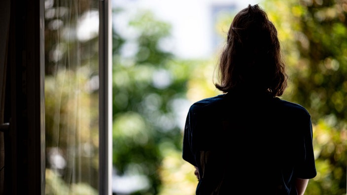 Eine Frau steht in ihrer Wohnung an einem Fenster und blickt nach draußen. 