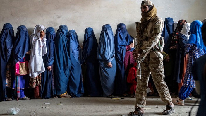 Bewacht von einem Taliban-Soldaten warten verschleierte Frauen in Afghanistan in einer Schlange auf Lebensmittelrationen.