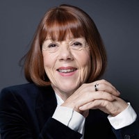 Die Porträtaufnahme von Gisèle Pelicot zeigt sie mit rotbraunem Haar und einem freundlichen Lächeln, die Ellbogen auf dem Tisch abgestützt und die Hände gefaltet, in einem edlen schwarzen Sakko.
