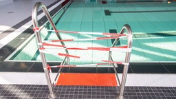 Die Leiter zum Schwimmbecken eines Hallenbads ist mit einem rot-weißen Flatterband gesperrt.