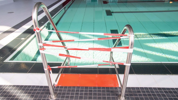Die Leiter zum Schwimmbecken eines Hallenbads ist mit einem rot-weißen Flatterband gesperrt.