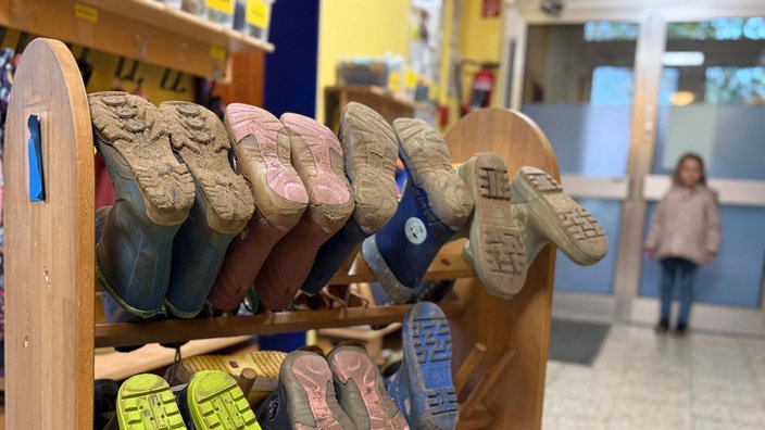 Ein Schuhregal auf Rädern, in dem Gummistiefel auf Stangen aufeinandergereit sind, steht im Flur eines Kindergartens. Dahinter steht ein Mädchen am Eingang der Kita.