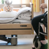Ein Bewohner sitzt in einem Altenheim in seinem Zimmer in einem Rollstuhl. Auf dem gemachten Bett liegt ein Stofftier.