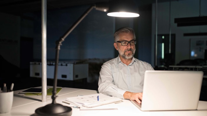 Ein mittelalter Mann mit Bart und Brille sitzt am Abend im Büro und arbeitet an seinem Notebook. 