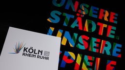 Das Logo der Olympia-Bewerbung "Köln Rhein Ruhr" auf einem weißen Blatt – im Hintergrund das zugehörige, vielfarbig gestaltete Motto: "Unsere Städte, unsere Spiele".