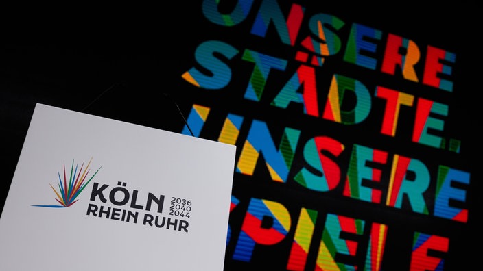 Das Logo der Olympia-Bewerbung "Köln Rhein Ruhr" auf einem weißen Blatt – im Hintergrund das zugehörige, vielfarbig gestaltete Motto: "Unsere Städte, unsere Spiele".