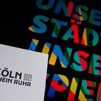 Das Logo der Olympia-Bewerbung "Köln Rhein Ruhr" auf einem weißen Blatt – im Hintergrund das zugehörige, vielfarbig gestaltete Motto: "Unsere Städte, unsere Spiele".