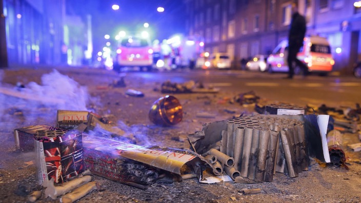 Abgebrannte Böller liegen nach der Silvesternacht auf der Straße, während im Hintergrund Einsatzkräfte der Feuerwehr stehen. 