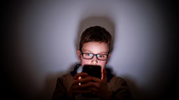 Ein Junge sitzt im Dunkeln und starrt gebannt auf den hellen Bildschirm seines Smartphones.