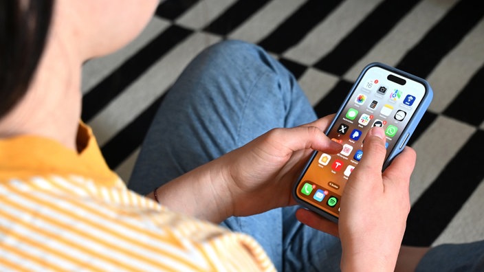 Nahaufnahme mit Fokus auf ein Smartphone, das verschiedene Apps anzeigt und von einer jungen Person gehalten wird.