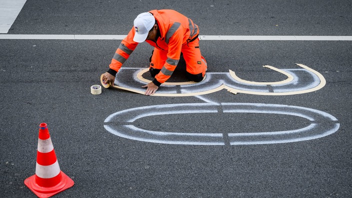 Ein Mann in orangener Arbeitsuniform klebt auf einer Straße mit Klebeband die Zahl "30" auf, um sie anschließend dort aufmalen zu können.