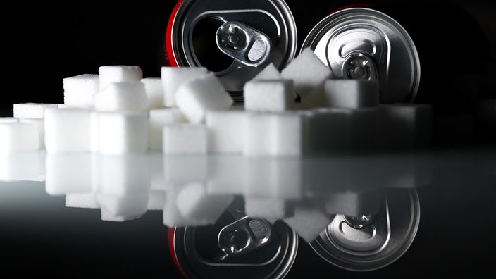Zwei Getränkedosen mit Softdrinks und zahlreiche Zuckerwürfel liegen auf einem Tisch.
