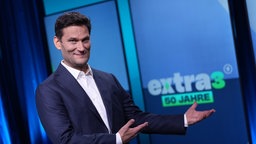 Christian Ehring, Moderator von "Extra 3", steht im Studio der Satiresendung, im Hintergrund steht "extra 3 , 50 Jahre", 15.09.2026. 