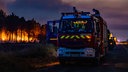 Feuerwagen stehen auf einer Straße in Saint-Jean-D'illac, im Hintergrund lodern die Flammen eines Waldbrandes. 