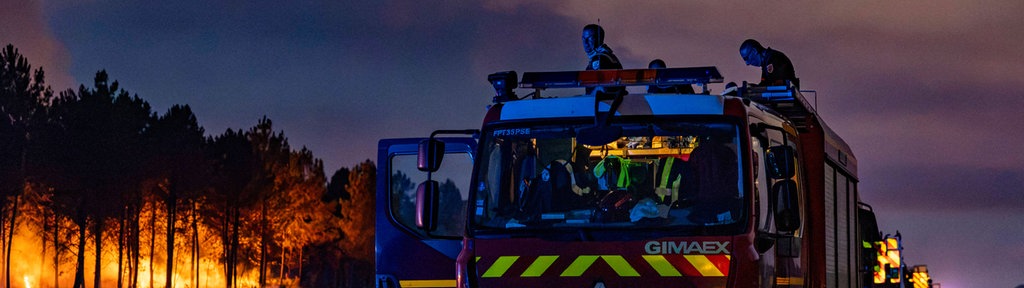 Feuerwagen stehen auf einer Straße in Saint-Jean-D'illac, im Hintergrund lodern die Flammen eines Waldbrandes. 
