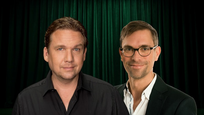 Die Kabarettisten Matthias Brodowy und Ingo Börchers stehen lächelnd vor einem dunkelgrünen Vorhang