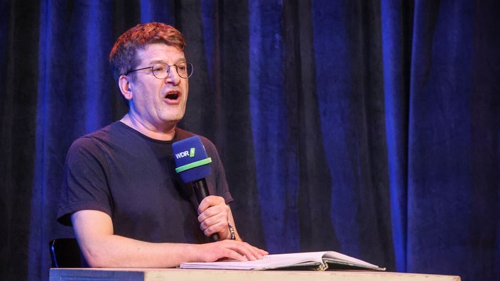 Nils Heinrich sitzt an einem Tisch mit einem WDR 5 Mikrofon in der Hand und liest auf der Bühne vom Theater am Dom