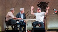 Das Kabarett-Trio "Wir Staatskünstler" sitzt beim Salzburger Stier 2026 an einem Tisch auf der Bühne