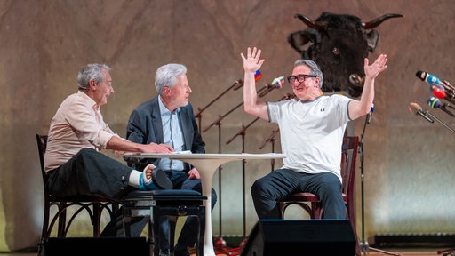 Das Kabarett-Trio "Wir Staatskünstler" sitzt beim Salzburger Stier 2026 an einem Tisch auf der Bühne
