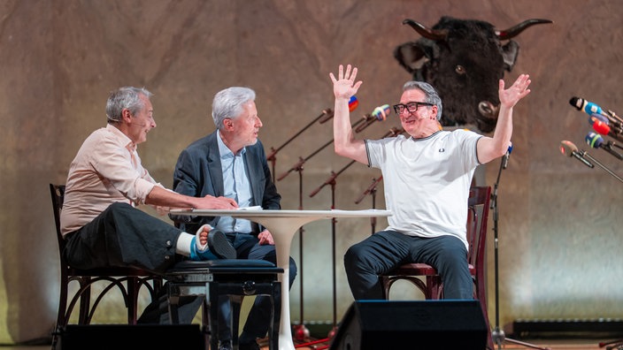 Das Kabarett-Trio "Wir Staatskünstler" sitzt beim Salzburger Stier 2026 an einem Tisch auf der Bühne