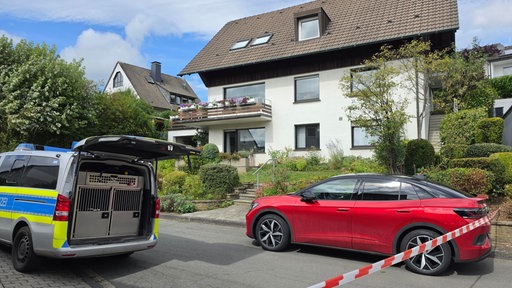 Ein Polizeiwagen und ein rotes Auto stehen hinter einer Polizeiabsperrung an einem Wohnhaus