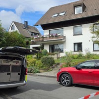 Ein Polizeiwagen und ein rotes Auto stehen hinter einer Polizeiabsperrung an einem Wohnhaus