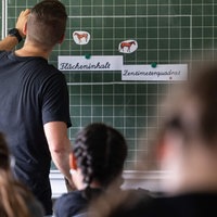 Ein Lehrer schreibt an eine Tafel, Schüler sitzen vor ihm im Raum