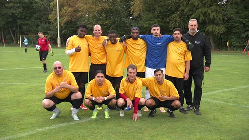 Eine Fußballmannschaft steht zusammen. Sie knieen oder stehen nebeneinader und legen die Arme über die Schultern des Nachbarn.