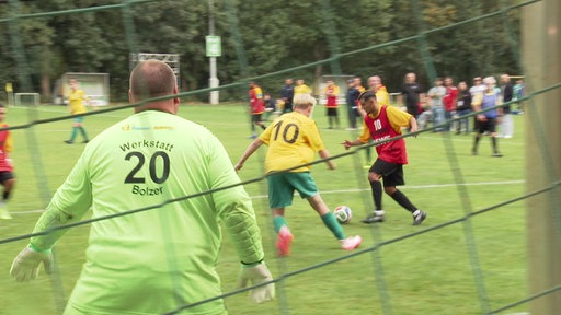 Zwei Fußballspieler spielen mit einem Ball in Richtung Tor. Der Torwart ist von hinten zu sehen. Im Vordergrnd ist das Tornetz.