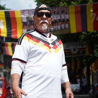 Achim Klimmeck in einem Deutschland‑Fußballtrikot, schwarzer Kappe und Sonnenbrille. Im Hintergrund hängen dutzende Deutschlandflaggen und ein Mannschaftsbild.