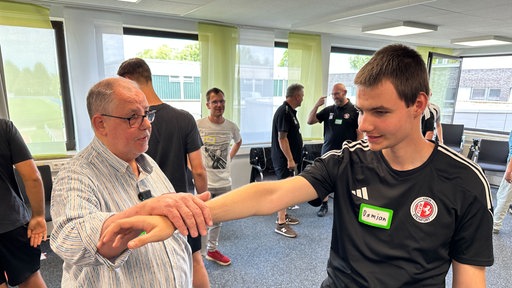 Ein junger Mann im Sporttrikot streckt seinen rechten Arm nach vorne, ein zweiter Mann greift mit seiner rechten Hand nach dem Arm des anderen.