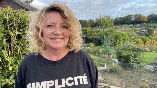 Eine Frau mit blonden Locken und Brille steht in einem Kleingarten
