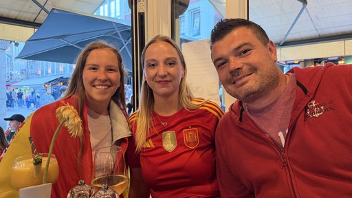 Zwei Frauen und eine Mann in Spanien-Trikots stehen vor einem Tisch und lächeln in die Kamera